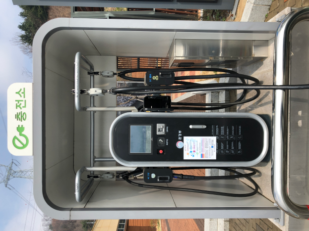 Eine Ladestation für Elektrofahrzeuge mit einer Maschine, Kabeln und einer Texttafel, vor einer Kulisse aus Gebäuden, Bäumen, Masten, Kabeln und dem Himmel.