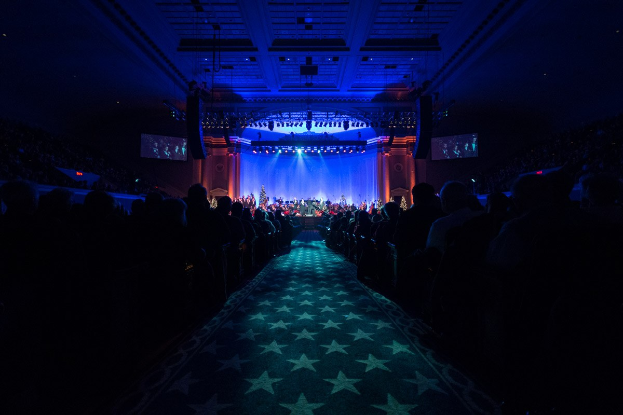 Ein großer Auditorium mit Sitzenden, eine Bühne mit Musikern und eine Decke mit Lichtern und Fernsehern, was auf einen Konzertsaal für die Nationale Convention 2019 hindeutet.