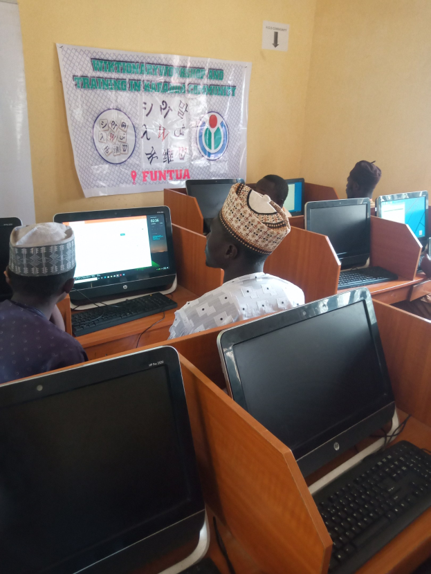 Gruppe von Menschen, die an Tischen mit Computerbildschirmen, Tastaturen und Mäusen sitzen und an einem Workshop zur Computertechnologie teilnehmen, mit einem Plakat im Hintergrund.