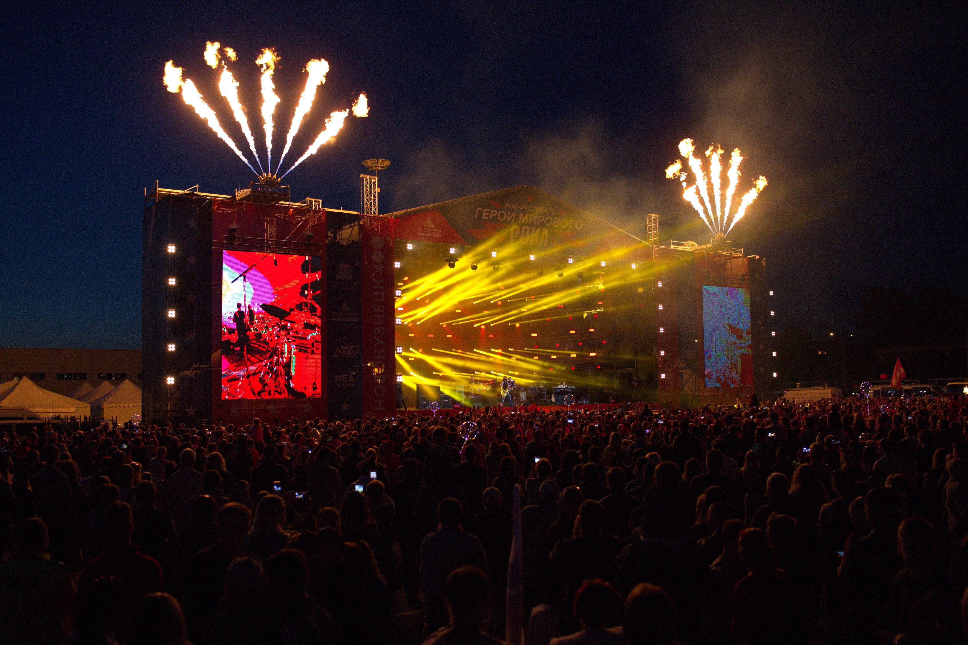 Eine große Menschenmenge auf einem Musikfestival schaut Performern auf einer hell erleuchteten Bühne zu, mit Zelten links und Feuerwerk in einem dunklen Himmel dahinter.