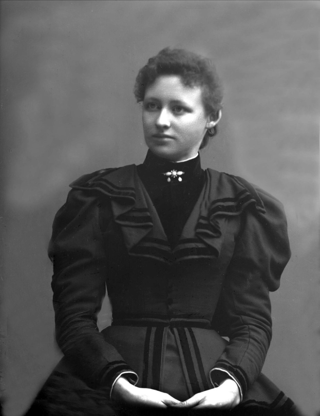 Ein Schwarz-Weiß-Foto einer Frau in einem eleganten, hochgeschlossenen Kleid mit langen Ärmeln, die selbstbewusst mit verschränkten Händen und einem ordentlichen Dutt steht.