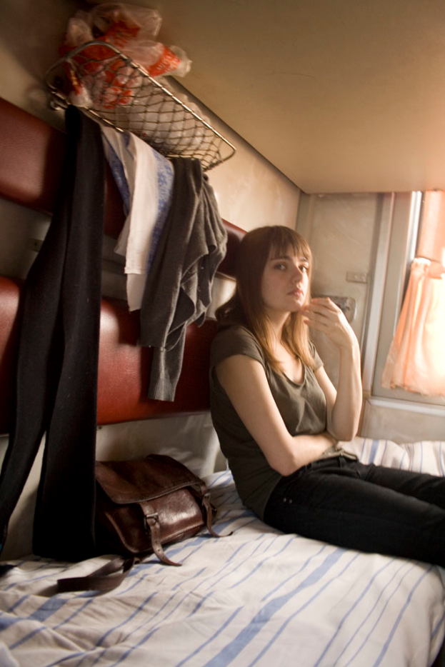 Eine Frau sitzt auf einem Bett mit einer Handtasche, Jacken an einem Ständer, einer Plastikabdeckung und einem Fenster mit Vorhängen.