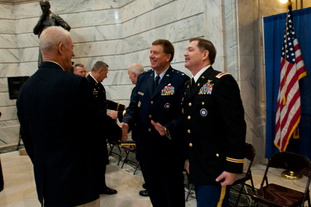 Menschen geben sich die Hand vor Stühlen, mit einer Statue, Flagge und Vorhang im Hintergrund.
