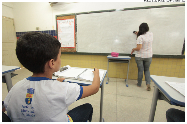 Ein Junge sitzt an einem Tisch und schreibt in ein Buch, während eine Frau die Tafel mit einem Putzlappen in einer Klasse mit mehreren Tischen, einem Plakat und einer Kiste auf einem Tisch abwischt.