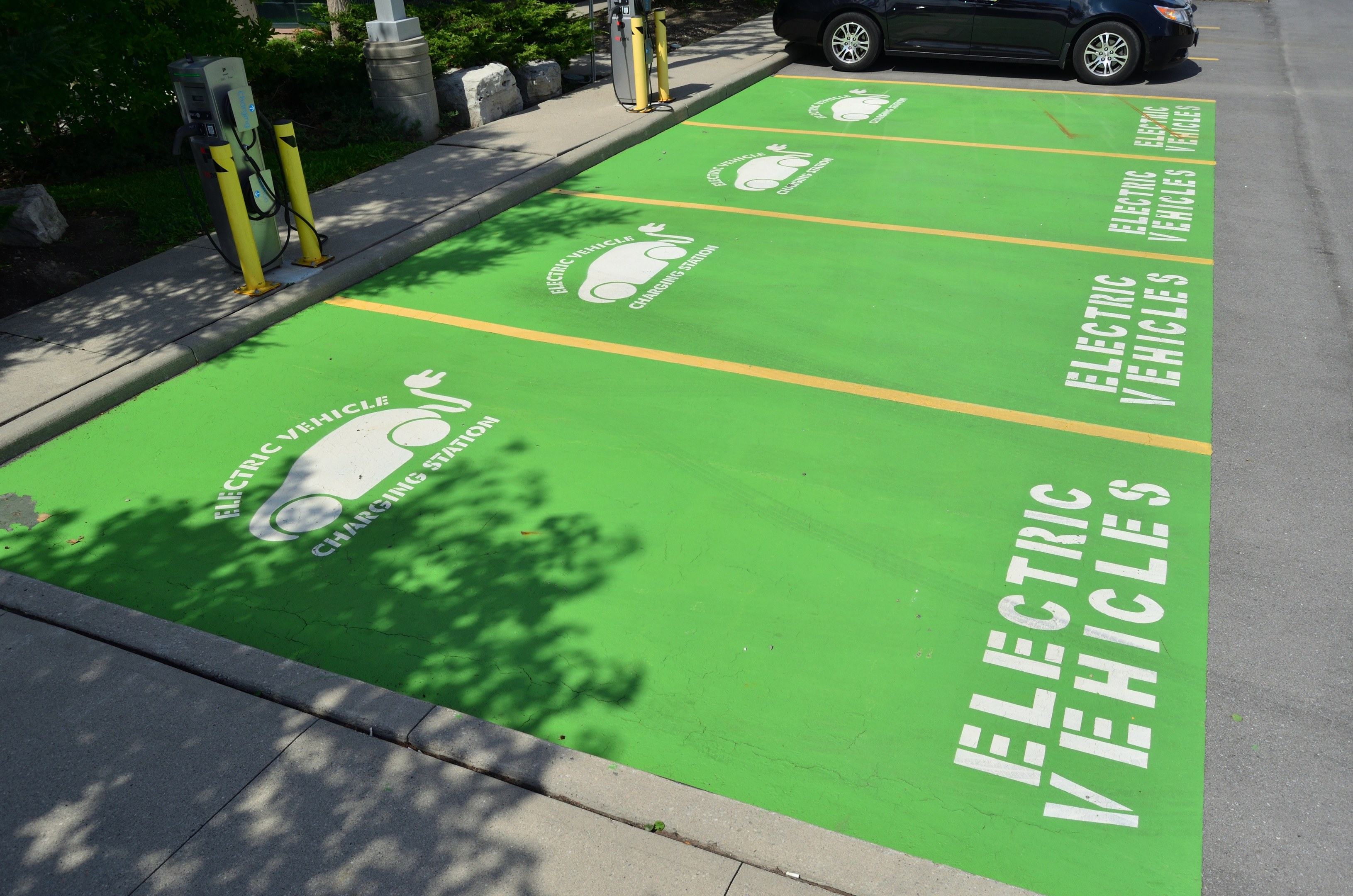 Elektroauto an einer Ladesäule mit grünem Bodenmatten mit Text und Bildern auf einem Parkplatz umgeben von Bäumen und einem Gebäude.
