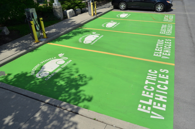 Elektroauto an einer Ladesäule mit grünem Bodenmatten mit Text und Bildern auf einem Parkplatz umgeben von Bäumen und einem Gebäude.
