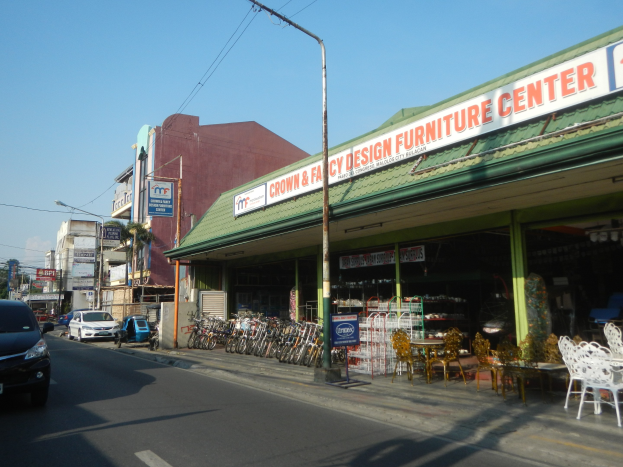 Eine belebte Stadtstraße mit fahrenden Fahrzeugen, parkenden Fahrrädern, Gebäuden mit Namensschildern, Laternen und Strommasten mit Drähten; ein Möbelgeschäft im Vordergrund und der Himmel im Hintergrund.