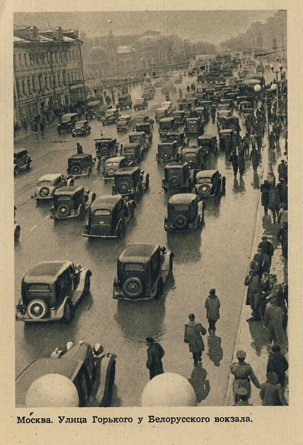 Ein altes Schwarz-Weiß-Foto einer belebten Straße mit Autos, Menschen und beleuchteten Gebäuden im Hintergrund, mit Text unten.