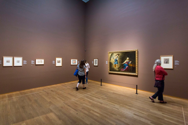 Eine Gruppe von Menschen geht durch eine Kunstgalerie mit gerahmten Fotos an den Wänden und einer beleuchteten Decke und trägt Taschen.