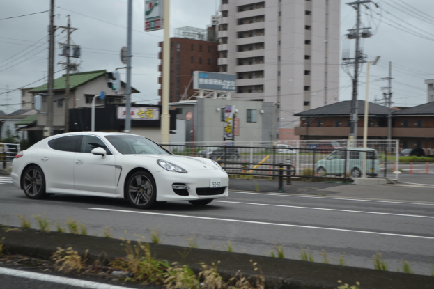 Ein weißer Porsche Panamera fährt eine Straße mit hohen Gebäuden, Strommasten, Laternen und Texttafeln entlang, mit einem Van und einem Auto im Vordergrund und Pflanzen unten, unter einem sichtbaren Himmel.