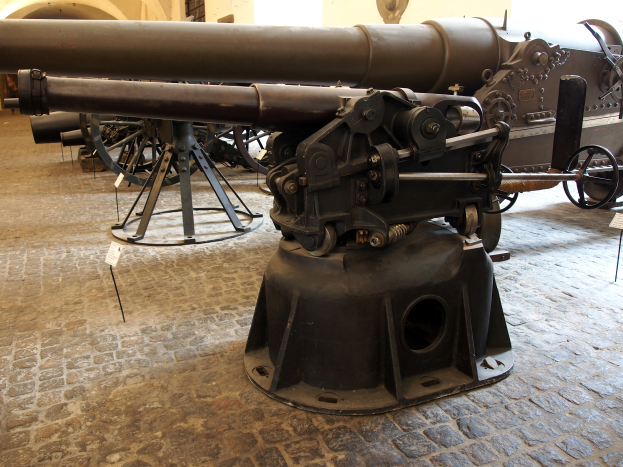 Eine große Gatling-Kanone mit einem langen Lauf und einem Seitengriff ist in einem Museum ausgestellt, umgeben von Bodenständen, mit einer fensterumgebenen Wand im Hintergrund, die an eine Burg erinnert.