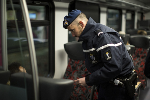 Ein Polizist in Uniform und Mütze steht in einem Zug und schaut auf sein Handy, umgeben von einigen sitzenden Passagieren und einer weiteren Person auf der rechten Seite.