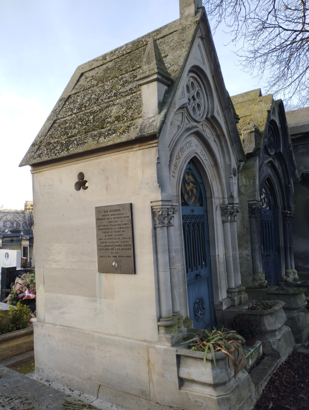 Eine kleine Kirche mit einer blauen Tür in einem Friedhof, umgeben von Gebäuden und Bäumen unter einem klaren blauen Himmel, mit einer Gedenktafel an ihrer Wand.