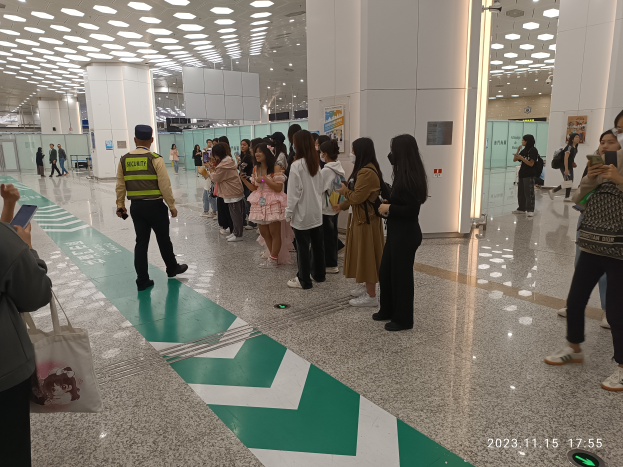 Eine Gruppe von Menschen steht in einer Reihe an einem Flughafen-Sicherheits-Check-in, einige halten Taschen und Mobiltelefone, mit Säulen im Hintergrund und Lampen an der Decke und einem Wasserzeichen am unteren Bildrand.