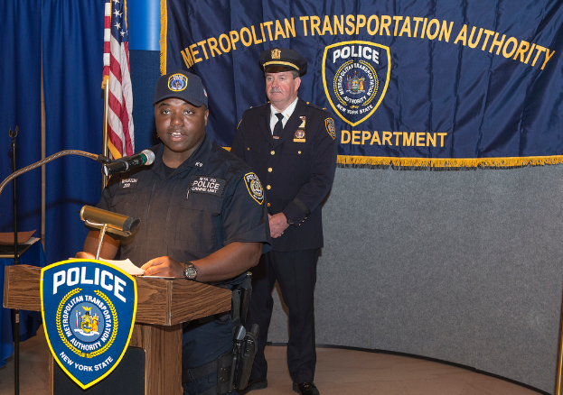 Zwei Polizeibeamte stehen vor einem Rednerpult, einer h√§lt Papiere und spricht in ein Mikrofon, mit einer Fahne, einem Banner und einem Tisch in der N√§he, und einer Tafel mit Symbolen und Text im Hintergrund.