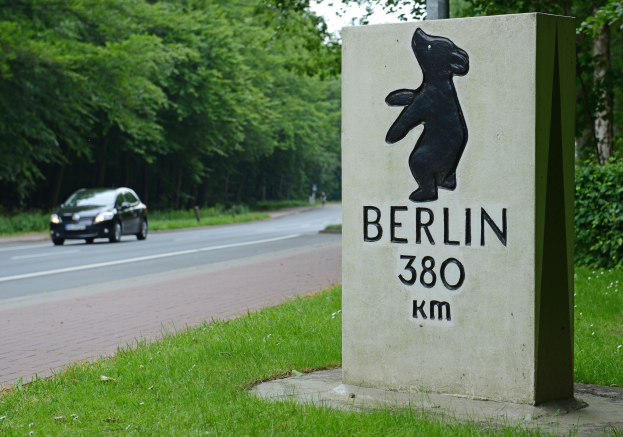 Ein Auto fährt auf einer Straße mit einem Schild, auf dem "Berlin 380 km" steht, auf der linken Seite, mit Pflanzen und Bäumen auf der rechten Seite und mehr Bäumen und einem klaren blauen Himmel im Hintergrund.