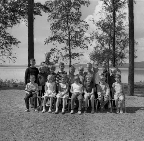 Schwarz-weiß-Foto einer Gruppe von Kindern, die auf Stühlen sitzen und vor einem See stehen, mit Bäumen, Pflanzen, Hügeln und einem bewölkten Himmel im Hintergrund, beschriftet mit "1940er Kinder am See."