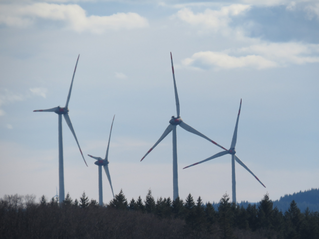 Eine Gruppe von Windrädern auf einem Feld mit Bäumen, Hügeln und Wolken im Hintergrund.