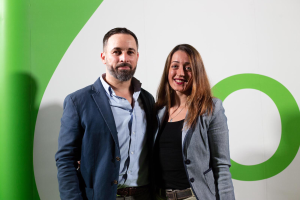 Ein Mann und eine Frau stehen nebeneinander, lächeln, vor einer grünen und weißen Wand in einem professionellen Setting.