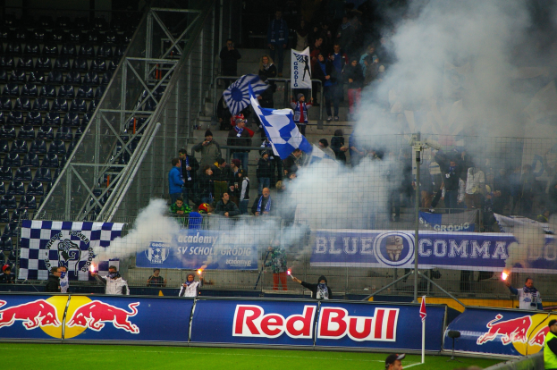 Menschen auf einem Fußballfeld in einem Stadion stehen, halten Fahnen und Schilder und Rauch steigt vom Boden auf, während leere Stühle links zu sehen sind.