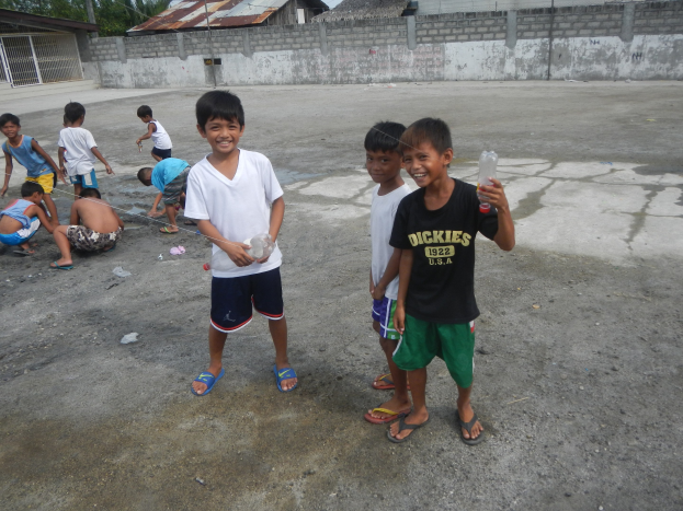 Junge Jungs, die in einer Parklücke stehen und Flaschen halten, mit Häusern, Bäumen und Polen im Hintergrund unter einem klaren blauen Himmel.