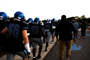 Eine Gruppe von Fussballspielern in Helmen und Stiefeln geht eine Strasse mit Bäumen und einem klaren blauen Himmel im Hintergrund entlang.