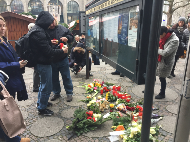 Menschen versammeln sich an einer Bushaltestelle mit Blumen und Kerzen auf dem Boden, einige halten Gegenstände oder eine Kamera, mit einem Gebäude, Bäumen und einer Scheune im Hintergrund.