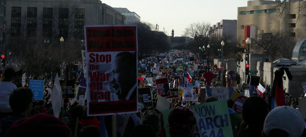 Eine Gruppe von Menschen, die auf einer Stra├če protestieren und Plakate halten, mit Geb├Ąuden, B├Ąumen, Laternenmasten und dem Himmel im Hintergrund.