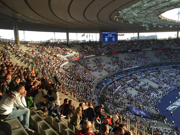 Große Menschenmenge in einem Stadion bei einem Fötballspiel mit einer Bühne rechts, Fahnen, Stangen, einem Bildschirm und der Allianz Arena in München, Deutschland im Hintergrund.