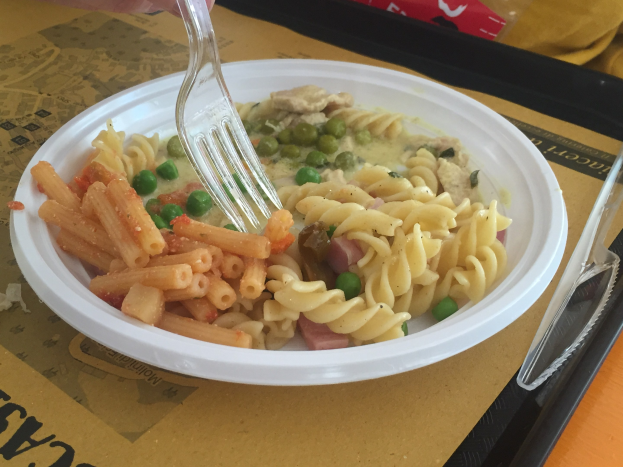 Eine Person hält eine Gabel über eine Schüssel mit Nudeln, Erbsen und Schinken, mit einem Tablett in der Nähe, auf dem ein Löffel und eine Abdeckung liegen.