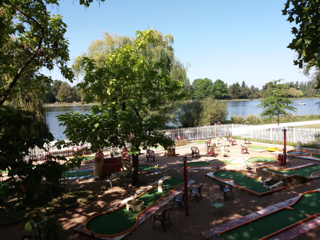 Minigolfanlage in einem Park mit grünem Gras, Bäumen und einem Zaun, umgeben von verstreuten Stühlen und Tischen, einem Gewässer mit einem Boot und einem sichtbaren Himmel.