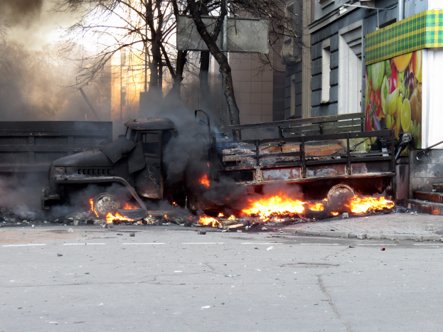 Ein Lkw brennt am Straßenrand, mit Rauch, der aus ihm aufsteigt, während im Hintergrund Bäume, Gebäude und ein klarer Himmel zu sehen sind.