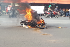 Ein Motorrad steht in Flammen am Straßenrand, mit einem Auto und einem Van im Hintergrund, umgeben von Menschen, einige tragen Helme, mit einem Banner und einem Zelt im Bild.