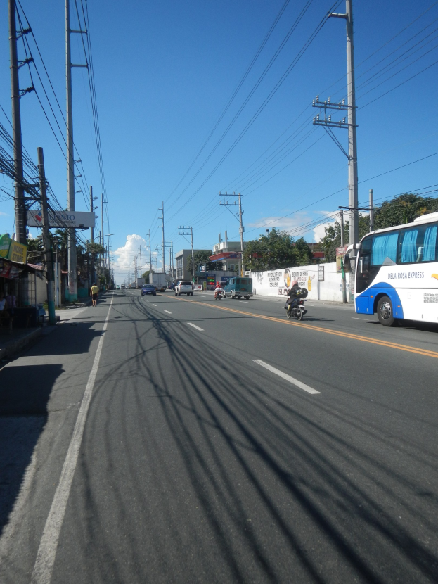 Ein Bus fährt auf einer Straße mit hohen Strommasten und Drähten, mit Geschäften und Passanten auf der linken Seite und einer Wand, Bäumen, Gebäuden und einem bewölkten Himmel im Hintergrund.