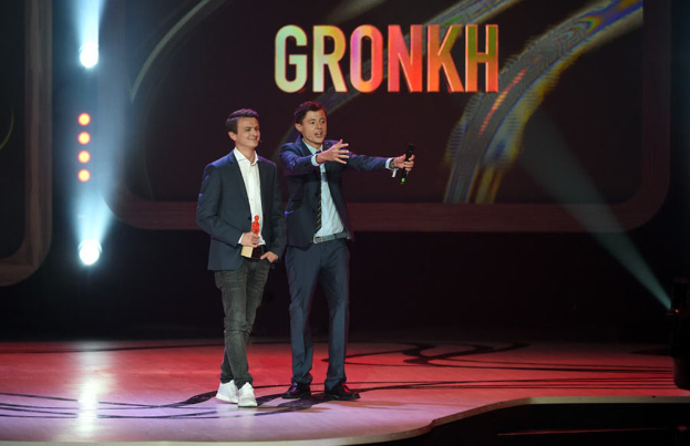 Eine Person in der Mitte mit einem Mikrofon sprechend und eine weitere Person links stehend und lachend vor einem Bildschirm mit Text und Lichtern.