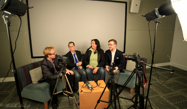 Eine Gruppe von drei Personen sitzt auf einem Sofa und gibt ein Interview, wobei eine Person in einem schwarzen Mantel und Hose links, eine Person in einem Mantel und blauer Krawatte mit einem Mikrofon in der Mitte und eine strahlende Person rechts zu sehen ist. Ein Mikrofonrecorder befindet sich auf dem Mitteltisch, Kameras sind auf der rechten und linken Seite und große Scheinwerfer sind vorhanden, mit einem Projektionsschirm im Hintergrund.