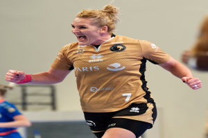 Eine Frau in braunem T-Shirt und schwarzen Shorts rennt auf einem Futsalplatz, feiert mit triumphierendem Gesichtsausdruck, während zwei andere Frauen im Hintergrund unscharf sind.