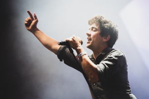 Ein Mann in schwarzem Hemd und Jeans singt leidenschaftlich in ein Mikrofon auf einer hell erleuchteten Bühne, während das Publikum im Hintergrund mitklatscht.