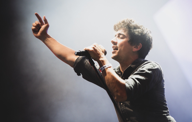 Ein Mann in schwarzem Hemd und Jeans singt leidenschaftlich in ein Mikrofon auf einer hell erleuchteten Bühne, während das Publikum im Hintergrund mitklatscht.