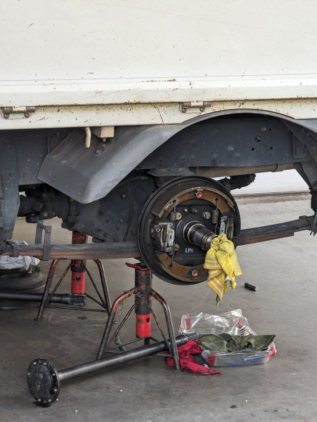 Ein gelbes Tuch liegt über einem Lkw, der in einer Garage geparkt ist, mit Werkzeugen und Gegenständen, die vor ihm auf dem Boden verstreut sind.