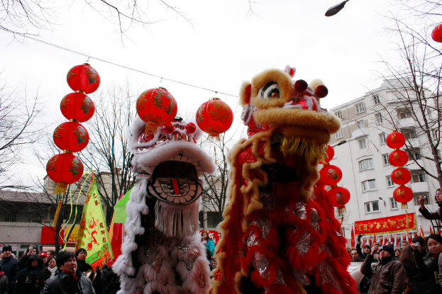 Puppen und Laternen an einem Haken mit einer Menge darunter, umgeben von Brettern, Gebäuden, Bäumen und einem erleuchteten Himmel.