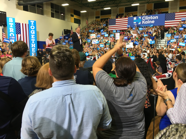 Eine große Menschenmenge steht vor einer Bühne, auf der ein Mann in ein Mikrofon spricht, eine Frau steht neben ihm, mit Flaggen, Transparenten und einem Lautsprecher auf der Bühne, und die Menge klatscht und jubelt.