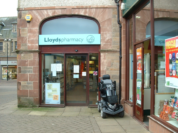 Eine Apotheke mit einem vor ihr geparkten Fahrzeug und einem Gebäude auf der linken Seite.