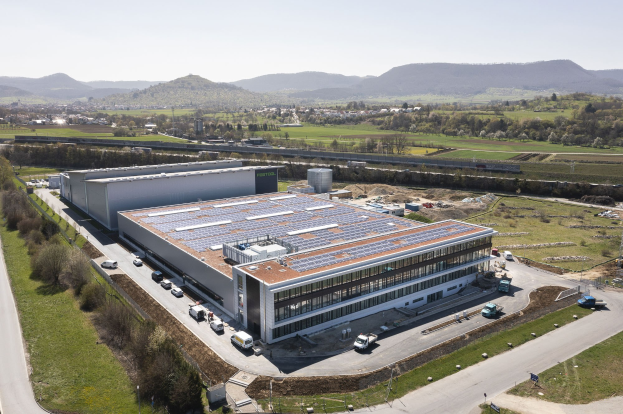 Luftaufnahme eines großen Gebäudes mit Solarpaneelen auf dem Dach, umgeben von Fahrzeugen, Bäumen, Gras und Polen, mit Hügeln und einem klaren blauen Himmel im Hintergrund.