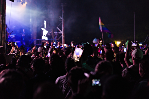 Menge von Menschen auf einem Konzert mit hochgehaltenen Handys, beleuchtet von Lichtern und umgeben von Pfählen und einer Flagge, während Musiker auf der Bühne spielen.