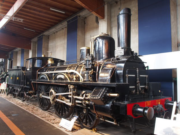 Alte Dampflokomotive in einem Museum ausgestellt, umgeben von Eisenbahnschienen und Informationsschildern, mit einer Wand und Deckenbeleuchtung im Hintergrund.