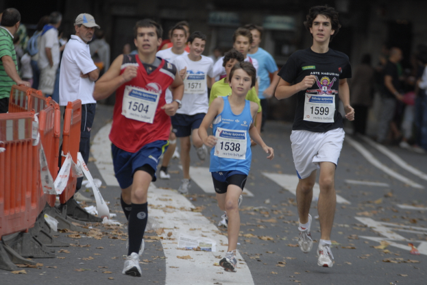 Läufer auf einer Straße mit Zuschauern auf der linken Seite hinter einem orangen Geländer, einige tragen nummerierte T-Shirts.