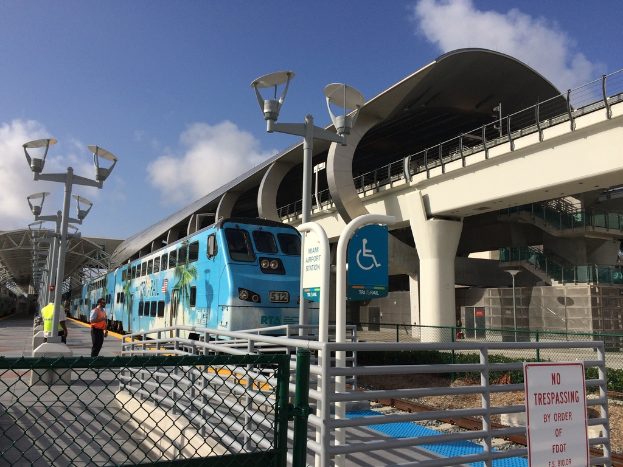 Mehrere Personen auf einem linken Bahnsteig mit einem Zug auf der rechten Seite, eine Brücke im Hintergrund, ein Zaun und einige Pfosten.