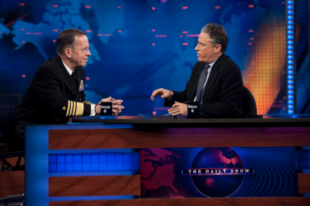 Zwei Manner in schwarzen Blazern, Hemden und Krawatten sitzen an einem Tisch mit Mikrofonen zwischen ihnen, vertieft in ein Gesprach vor einem Fernsehbildschirm und einem blauen Hintergrund.