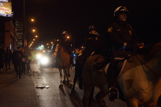 Drei Personen mit Helmen reiten nachts auf Pferden auf einer Straße, mit Fahrzeugen dahinter und Passanten auf einem nahegelegenen Fußweg, umgeben von Gebäuden, Polen und Bannern.